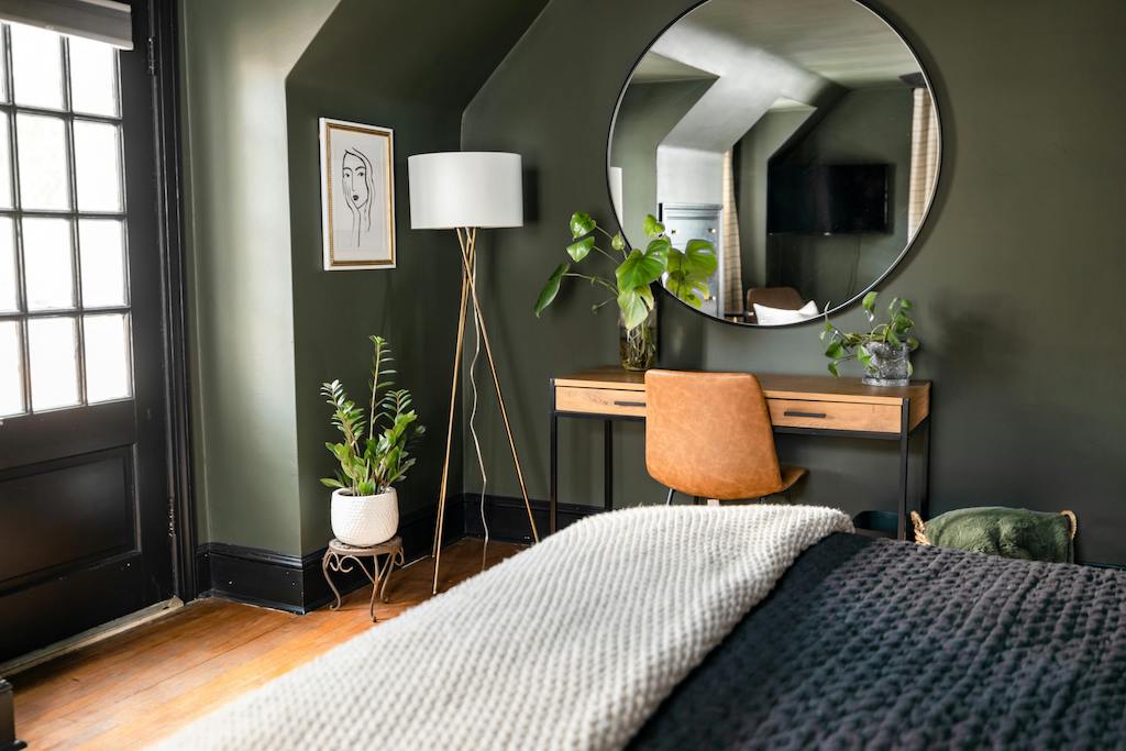 view of a corner of a bedroom with part of a bed in foreground and house plants, standing floor lamp and large round mirror on wall in background