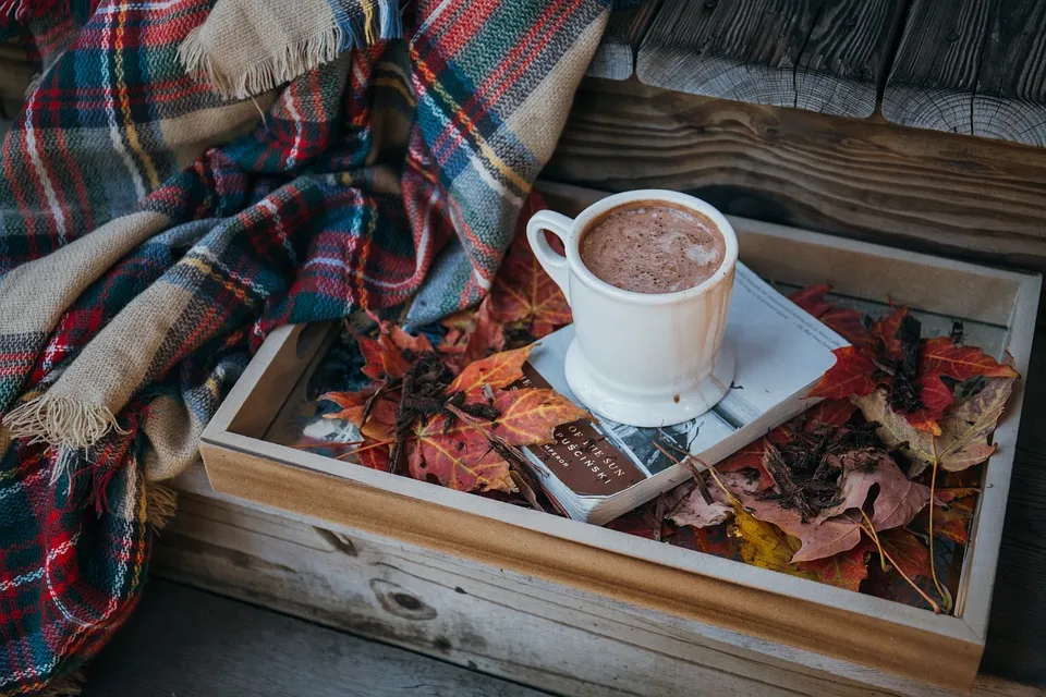 A chunky mug of hot chocolate on top of a book on wooden tray. Autumnal leaves scattered on the tray and a checked blanket nearby.