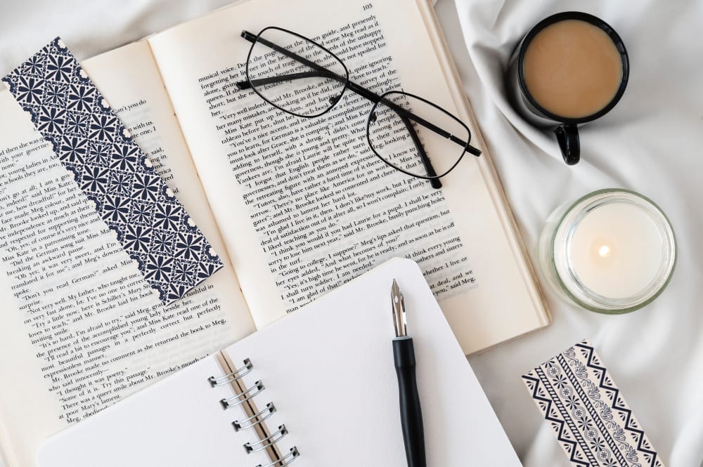 Image shows an open book with glasses and a book mark. On top is a blank open journal and a fountain pen