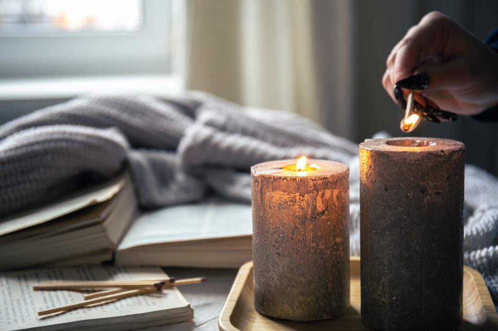 Image shows two candles side by side in the foreground with a cozy blanket and an open book in the background. 