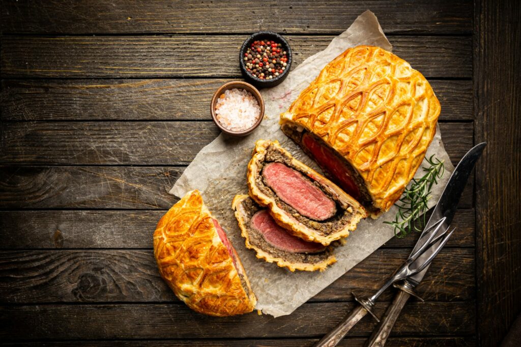 a beautifully encrusted beef wellington sliced with fork and knife in lower right corner and salt and pepper in small bowls at the upper left