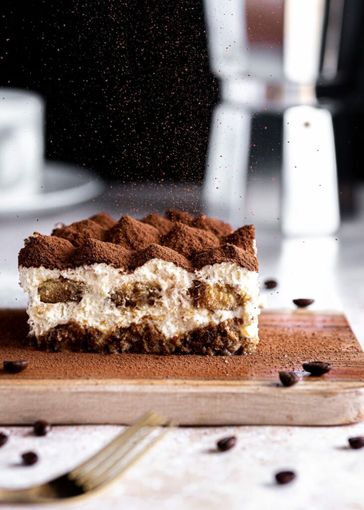 A slice of tiramisu on a wooden board with a dessert fork in the foreground and scattered coffee beans